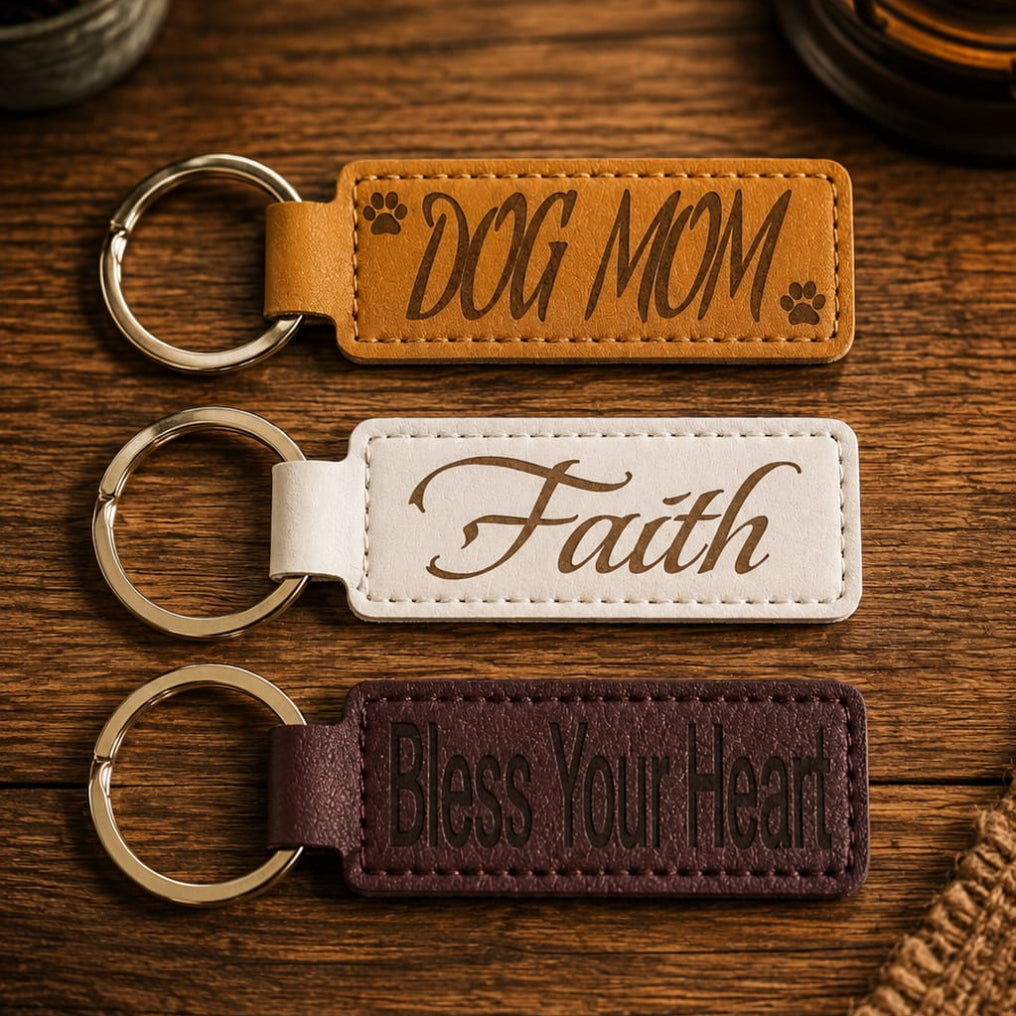 Three personalized leather keychains with metal key rings are arranged on a rustic wooden surface. The top keychain is tan with the words 'DOG MOM' and small paw prints; the middle is white with the word 'Faith' in cursive; the bottom is dark brown with the phrase 'Bless Your Heart' in dark lettering.